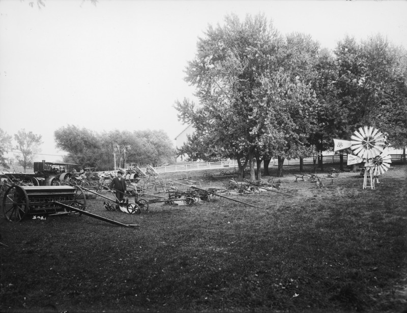 Drawn farm machinery lined up for exhibit. A front row contains windmill motors. A steam engine sits at the rear of the assembled machinery. A man stands in the second row with his hand on a two-bottom plow. There is a white fence in the rear of the machinery exhibit.