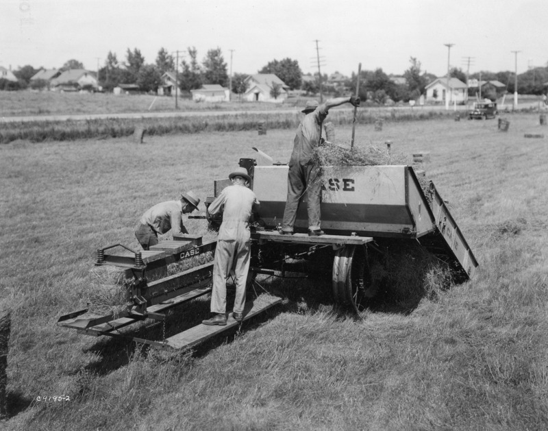 Three men working on a tractor-drawn Case hay baler in the foreground. A harvested hay field can be seen behind the baler. Residential housing across a gravel road is in the background.