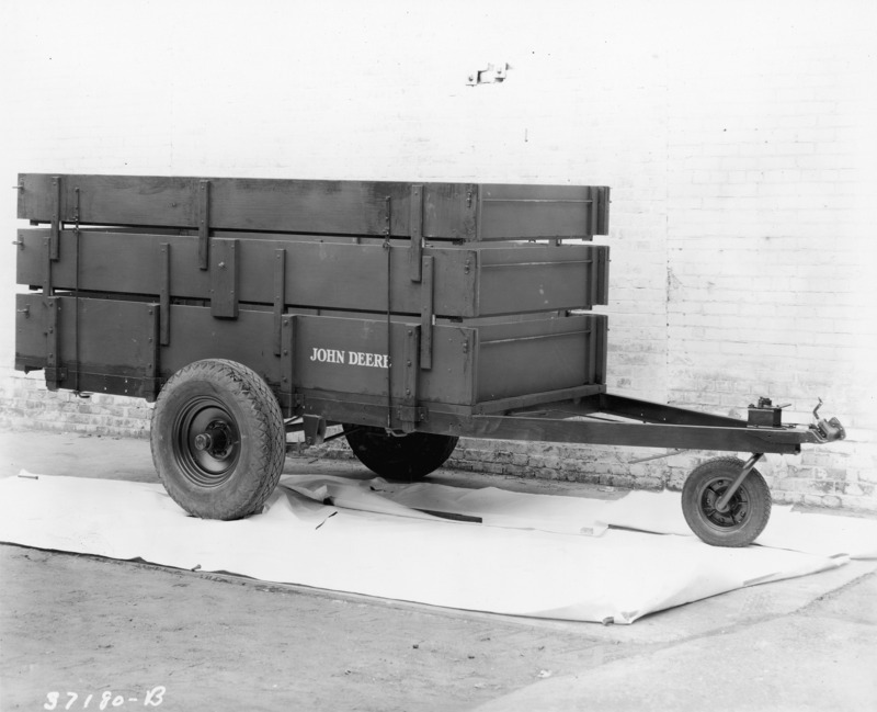 A John Deere farm freight trailer. The rubber-tired slat box trailer is standing on white paper or canvas against a painted brick wall.