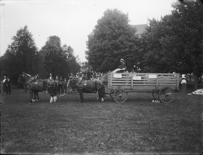 A four-horse team hitched to a hog wagon with one driver. Iowa State College (University) insignia on the harness of the horses are visible. Men and women in fine dress and looking at the horse team and wagon are on the far side. Behind all can be seen Old Botany (now Carrie Chapman Catt Hall) shrouded in trees. The photograph was taken in the fall of 1904, probably on Excursion Day.