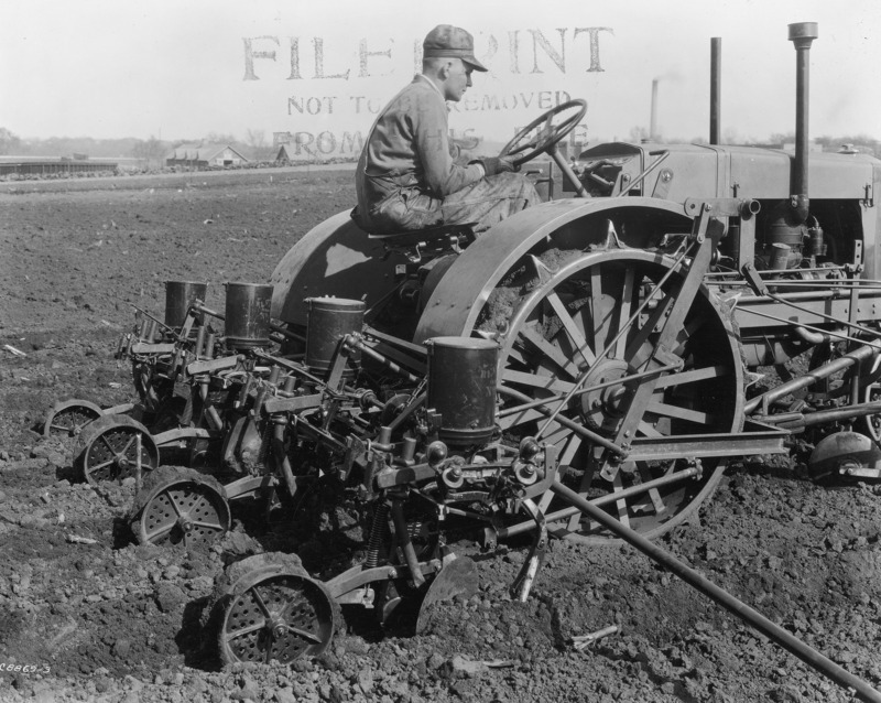 A four-row corn planter being pulled by a steel-wheeled tractor. A driver is at the wheel of the tractor. Buildings can be seen across a road in the distance. The photograph was taken April 27, 1931.