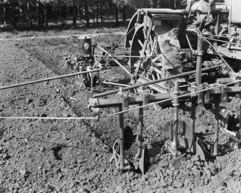 A four-row corn planter being pulled by a steel-wheeled tractor. The photograph is a close up of the part of the equipment which mounts on the front of the tractor. The photograph was taken April 27, 1931.