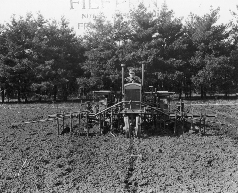 A four-row corn planter with front and rear components mounted on a steel-wheeled tractor. The photograph is from the front. The rear edge of the plowed field is directly behind the tractor. Along this boundary runs a line of conifer trees. A driver with a hat is seated on the tractor. The photograph was taken April 27, 1931.