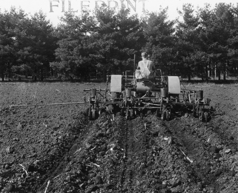 A four-row corn planter being pulled by a steel-wheeled tractor. The photograph is from the rear. A man in overalls and a hat is driving the tractor. The photograph was taken April 27, 1931.