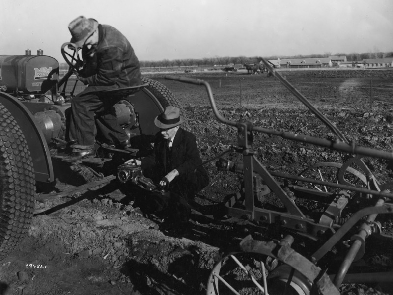 A tractor driver and Iowa State Professor J.B. Davidson looking at an Iowa integrating and recording traction dynamometer, mounted on the linkage between the tractor and the piece of farm machinery being pulled. The farm tractor has rubber tires. The machinery has iron wheels.