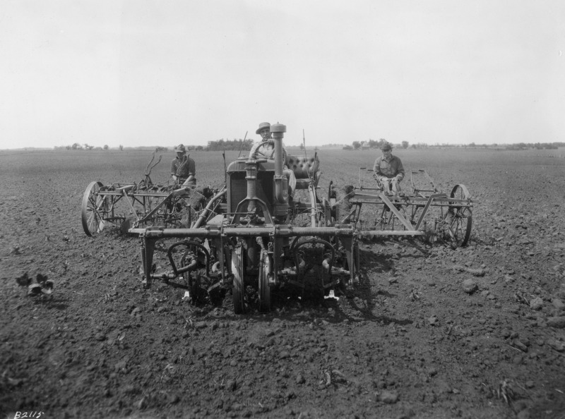 A tractor-mounted six-row cultivator seen from the front. One man is driving the tractor.One man is also seated on each of the two rear sections of the cultivator, one on each side of the machine. The machinery and the tractor have steel wheels. They are in the middle of a very large plowed field.