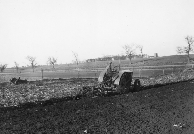 A field being plowed by a steel-wheeled tractor pulling a small plow. A man in work clothes is driving the tractor. On the horizon is a farmstead.