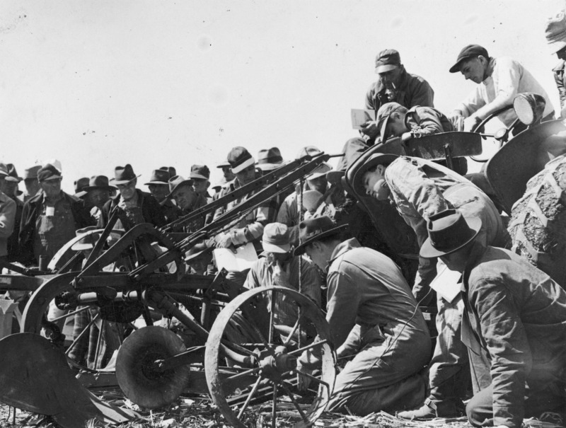 Dale Wollsoncroft demonstrating plow adjustment at a training event in State Center, Iowa, in the spring of 1949. A crowd of men is gathered around an instructor who is kneeling on the ground at the front end of a plow that is in turn connected to a tractor.