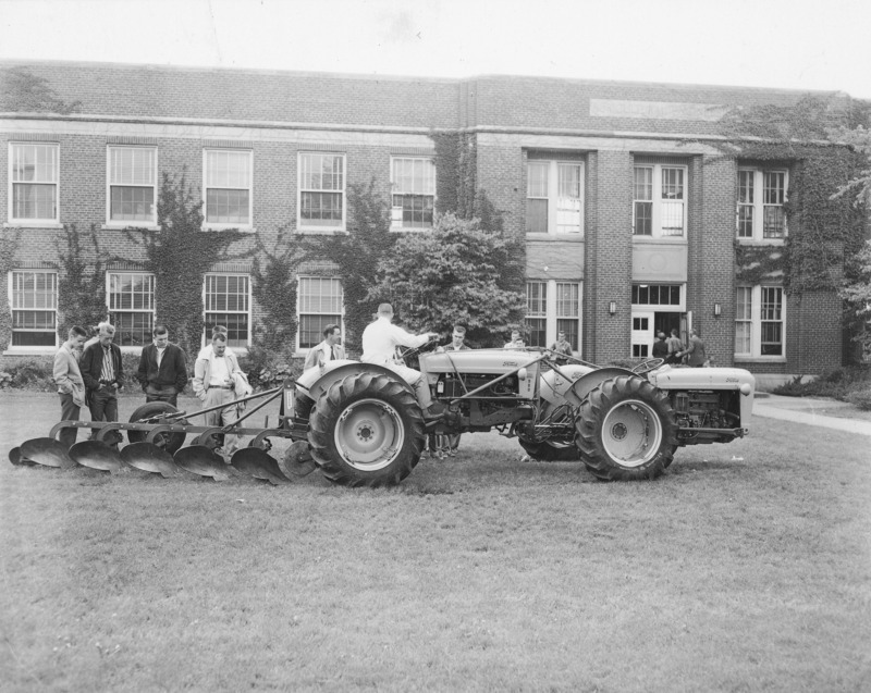 Two Ford tractors linked together as a single drivable unit and hitched to a five bottom plow. The machinery is sited in front of Davidson Hall. Ten men are looking at the machinery, another is sitting in the driving seat of the power unit. Another group of men is entering the front door of Davidson Hall, which is in the background.