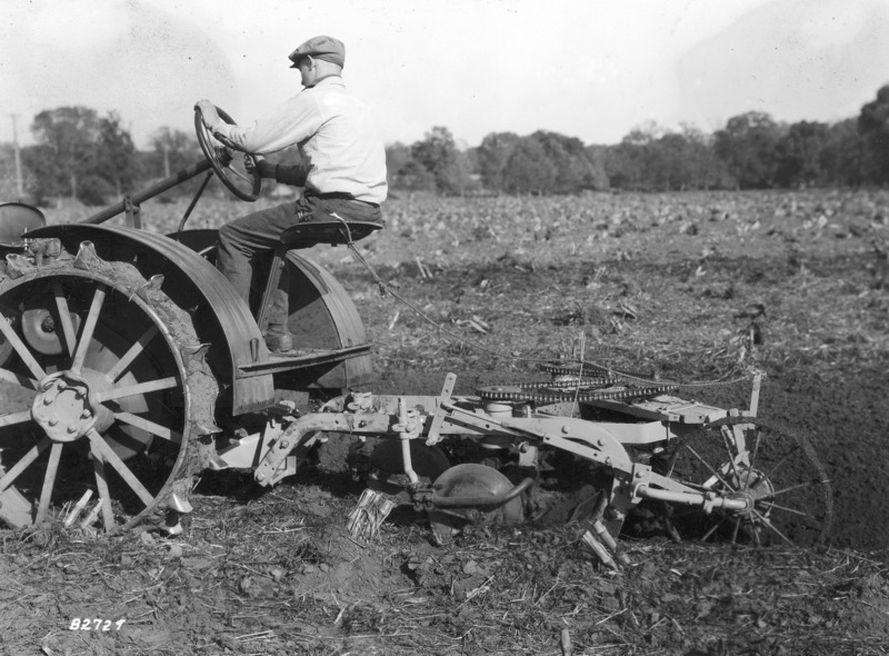 A driver on a steel-wheeled tractor pulling a plow with an unusual chain gearing system. A tree line in the background is the boundary for the field of corn stubble being plowed.