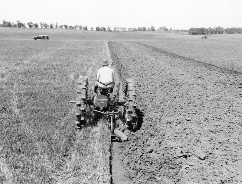 A driver on a steel-wheeled tractor pulling a plow toward the horizon through an unplowed field. Two automobiles are in the field in the far left distance. Another tractor is in the far right distance. A tree line on the horizon defines the edge of the large field.