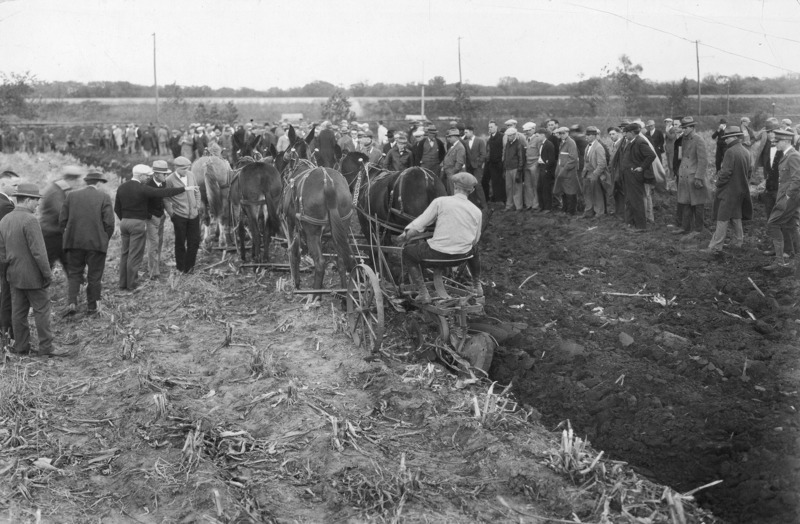 A demonstration of plowing with a six-horse and mule hitch, a technique that reduces side draft and increases efficiency. A large gathering of men are on both sides of the furrow as the driver plows ahead toward the end of the field on the image right.