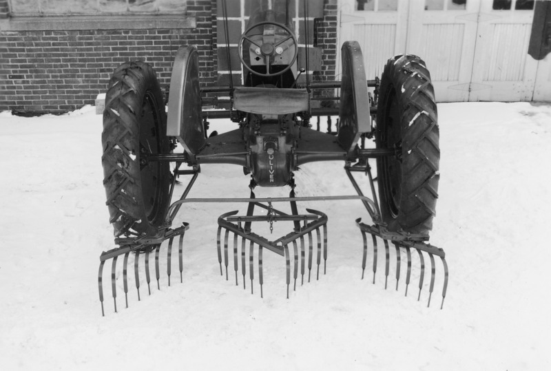 A rear view of an Oliver tractor with a spring tooth weeder rear attached for cultivation. However, the ground is snow covered. The tractor is parked close to a brick building.