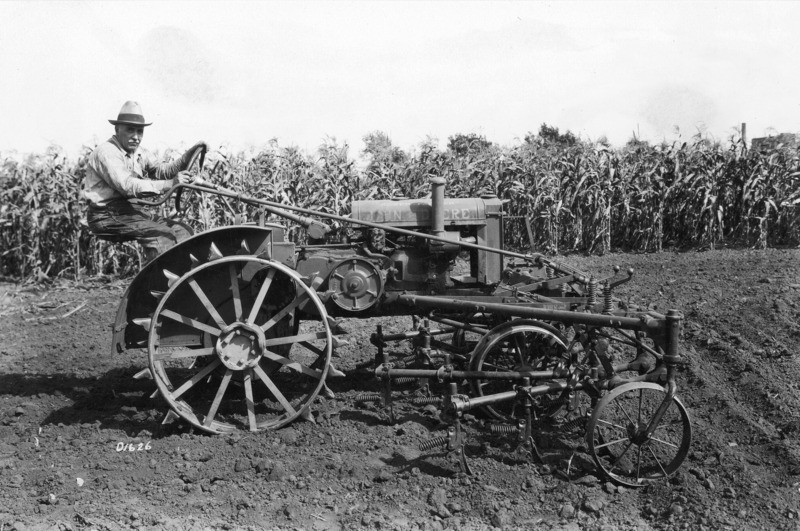 A steel-wheeled John Deere tractor with a cultivator mounted on the front. The driver on the stationary tractor is posing for the camera. In the background is a field of mature height corn.