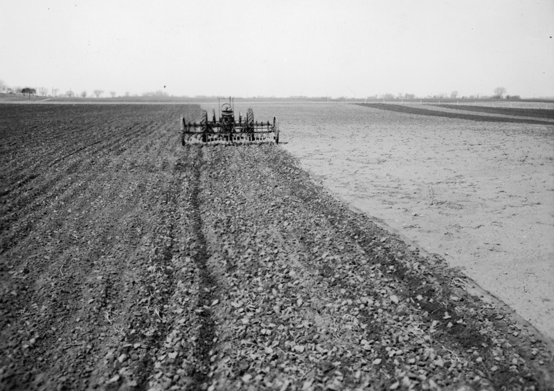 A rubber tired tractor with an attached field cultivator preparing a seedbed for corn. The tractor is stationary without a driver. It is viewed at middle distance from the rear, in the middle of a large field, untilled except for where this cultivator has been used.