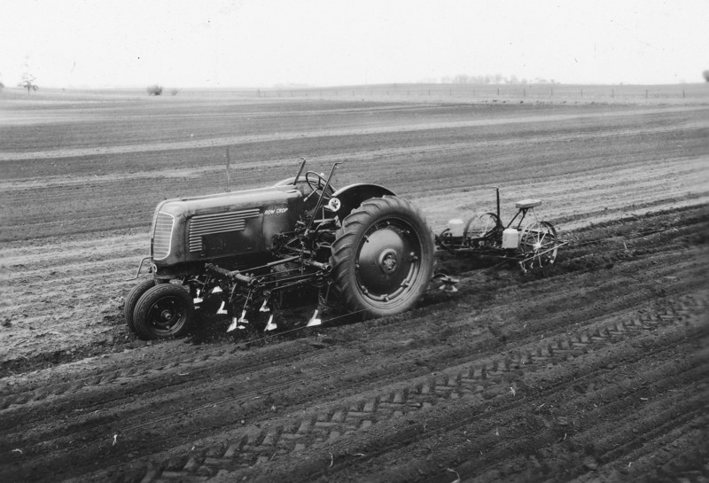 An Oliver Row Crop 70 tractor with a mounted cultivator and a two- hopper planter hitched behind. The set up is designed for one operation seedbed preparation and planting. The tractor is without a driver but stopped in mid-field. Cultivated fields stretch to the horizon.