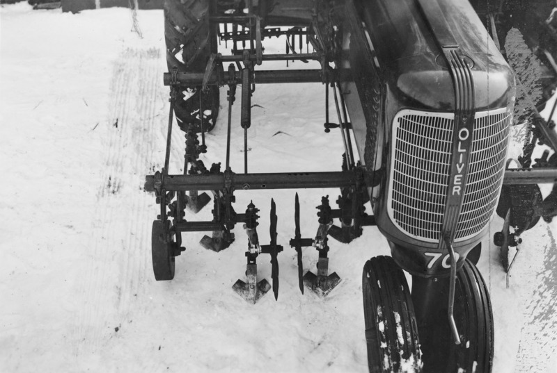 A close up view from above of a right section of a cultivator mounted on an Oliver tractor. The ground is snow covered.