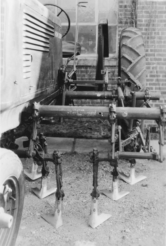 A close up view of the left front side section of a cultivator mounted on an Oliver tractor. The driverless tractor sits astride a sidewalk and backed against a brick building.