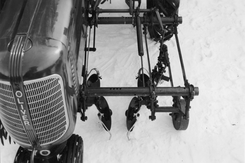 A rear view of an Oliver tractor with a spring tooth weeder rear attached for cultivating. However, the ground is snow covered. The tractor is parked head in against a brick building.