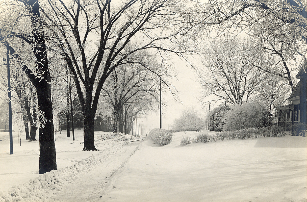 Photograph of a pathway cleared through the snow running east between the Farm House and Creamery, First (when it was probably known as the Farm Foreman’s Cottage) on the right (south side), and Landscape Architecture (when it was known as the Horse Barn) on the left (north side). Annotation: “Looking east Farm House on right, Horse Barn on left, after 1907 where the electric line turned north of Ag Hall & Margaret Hall.”