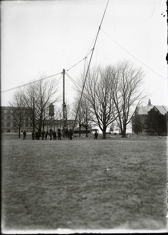 Photograph showing the raising of the flagpole on central campus. In the background Bearshear Hall, Marston Hall, the Marston Water Tower, the Hub, and Mirrill Hall can be seen. Annotation: "Erecting of steel flagpole, ca. March 1908. Beardshear Hall, Marston Hall, water tower, Hub, and Morrill Hall in background."