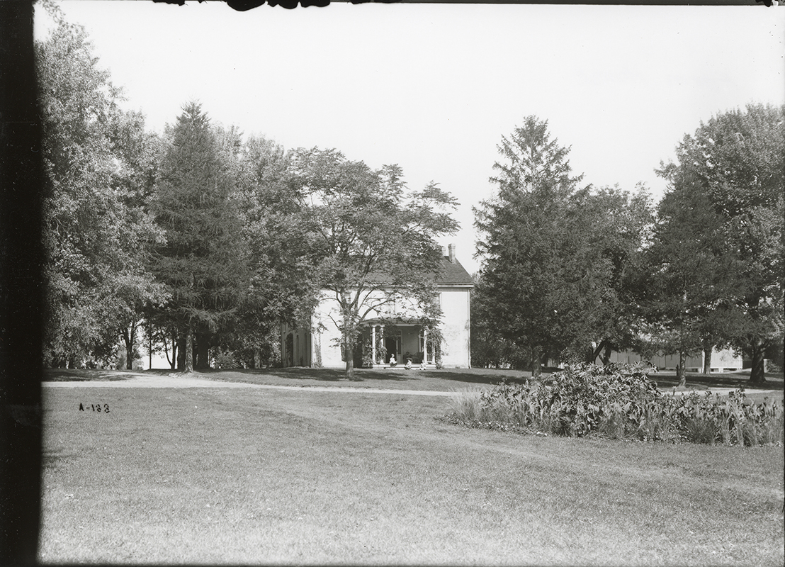 Photograph of the south side of the Farm House showing the front porch. The Cattle Barn, First can be seen in the background to the right (north-east) of the Farm House. Annotation: “Farm House. Cattle barns in background. Sept. 22, 1905 from south.”
