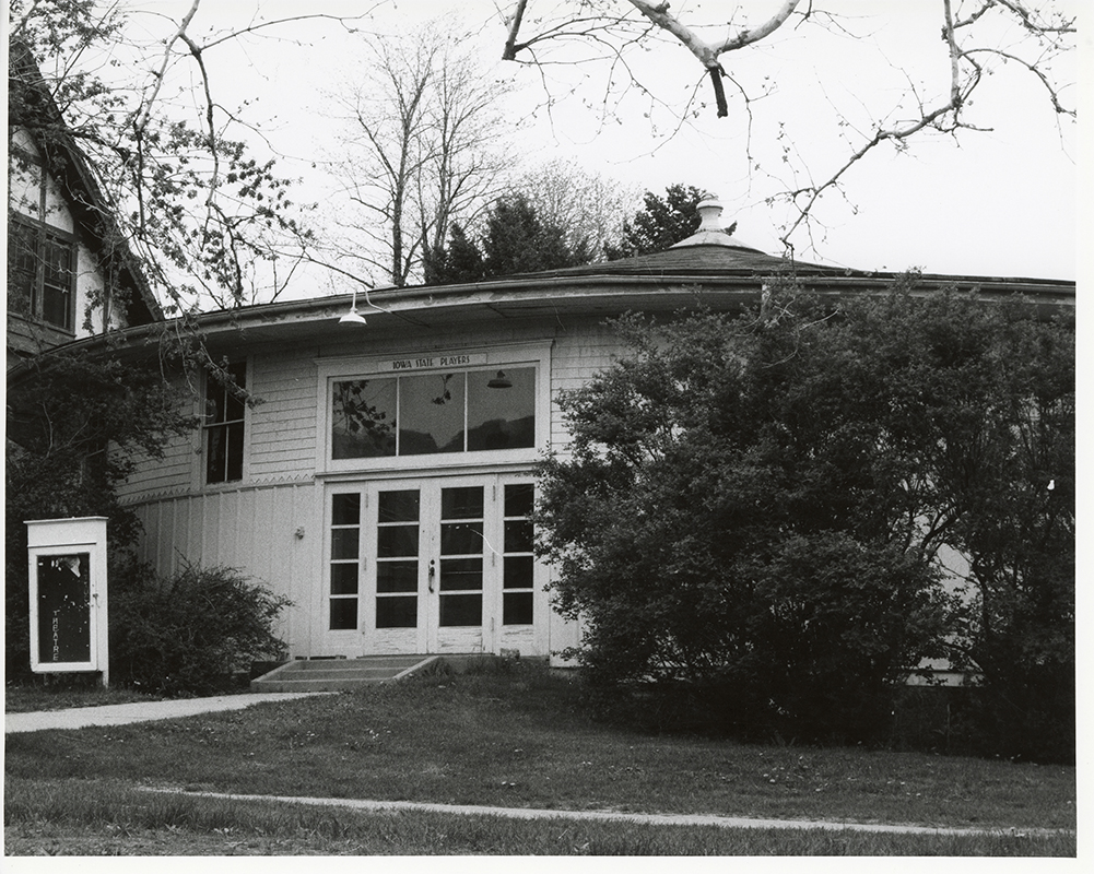 Photograph of the south door of Shattuck Theatre, facing north-northwest. There are bushes to the right of the door and a sign to the left.  