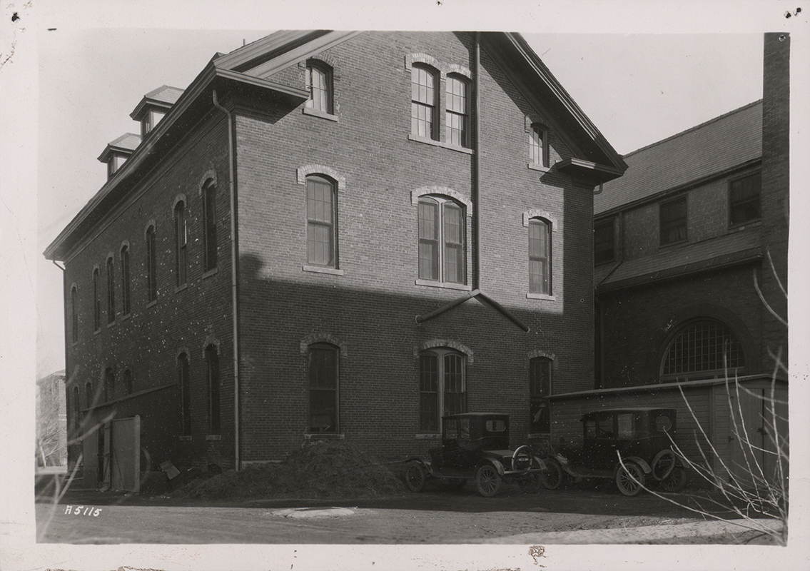 Photograph of North Hall on the Iowa State campus, showing the west and north elevations. The image depicts a three-story brick building with arched windows and adjacent structures, with early automobiles parked nearby. Annotation: North Hall 1896â€“1900 "Over Margaret Hall Kitchen."