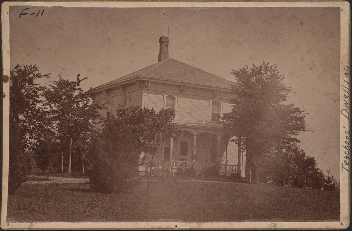 Photograph of Mortensen Cottage, shown from the front elevation. The structure features a central chimney, and a covered porch, surrounded by trees and landscaped grounds. Annotation: "Teachers' Dwelling."