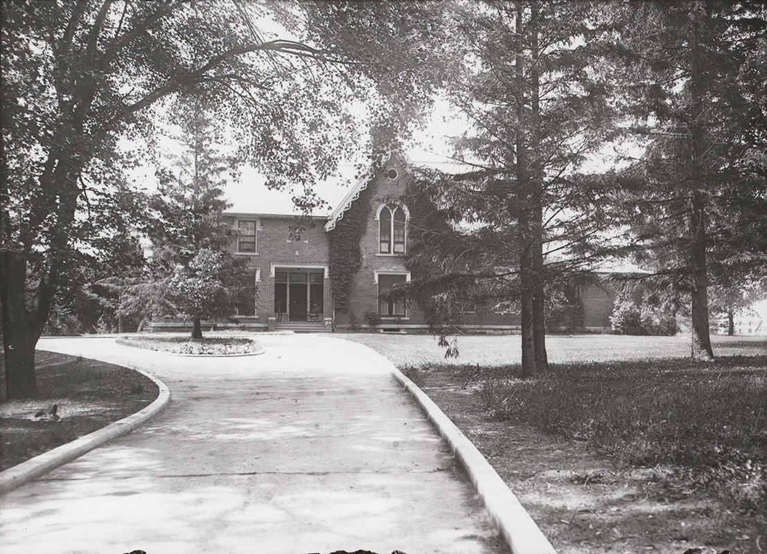 Photograph of Music Hall with surrounding landscape, showing the north elevation of the building. The image depicts a brick structure partially obscured by mature trees, with a curved driveway leading toward the entrance and landscaped grounds in the foreground. Annotation: "Old Music Hall. n.d. neg. 1310."