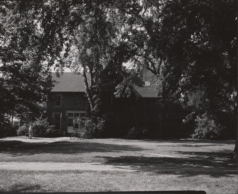 Photograph of Music Hall on the Iowa State campus, showing the north elevation. The image depicts a brick building partially obscured by mature trees, with shaded lawn in the foreground and a pathway leading toward an entrance. A person stands near the building, and a bicycle is visible nearby. Annotation: "Music Hall. 1954. neg 05120."