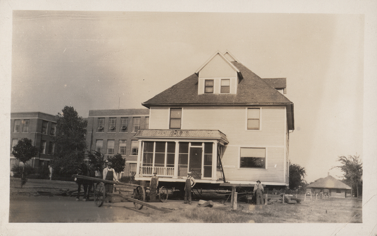 Photograph of Norton House being moved on the Iowa State campus. The image shows a two-story house elevated on supports, with workers and equipment positioned around the structure during relocation. The nearby campus buildings are visible in the background. Annotation: "Moving Coover Cottage which became Norton House. Norton House."