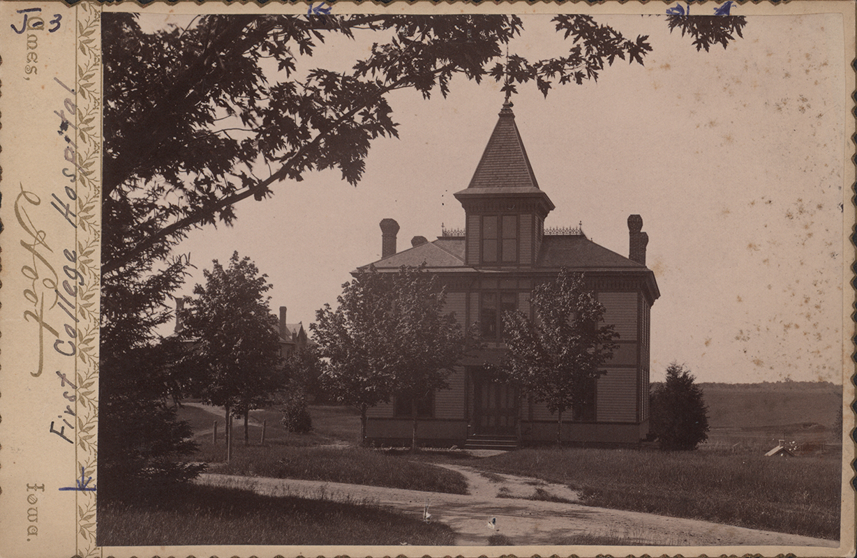 Photograph of the Sanitary Building, which served as the first college hospital. It is a two story building with a pointed cupola. There are trees directly in front of it on either side of the door.