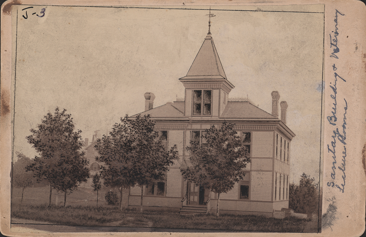 Photograph of the Sanitary Building, which also housed veterinary lecture rooms. It depicts a two story building with a square cupola. Three chimneys are visible on the roof, and three trees are directly in front of the building.