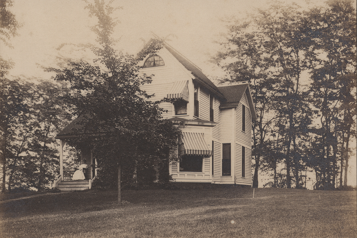 Photograph of Coburn House. It is a white two-story house with a tree in front. A woman in a white dress sits on the front porch.