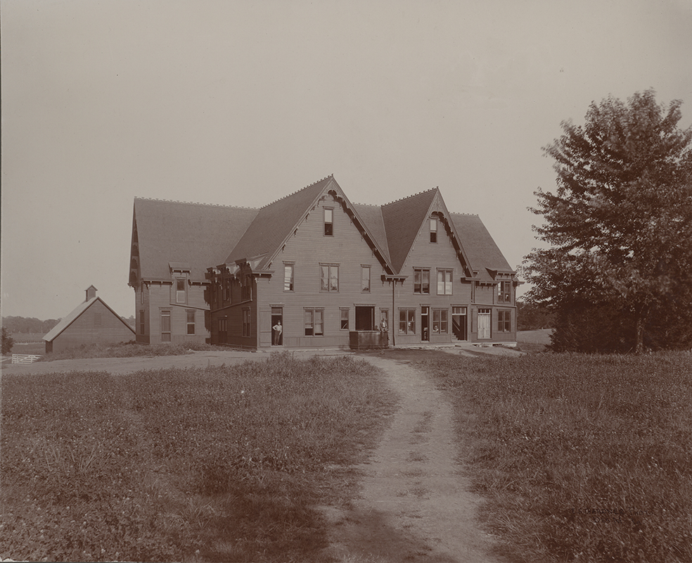 Photograph of the second creamery. It is a large three-story building. A barn sits in the backround to the left. Two men are outside the building: one leaning against a doorframe and another sitting on a box in front of a wider door. The caption reads "Creamery, 1892-1904 on site of East Hall."