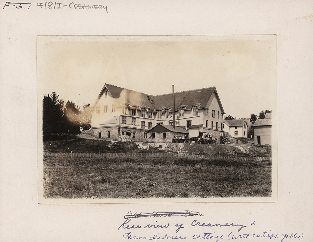 Photograph of the second creamery building from the rear, with the Farm Laborer's Cottage. The creamery is three stories, and a one-story building sits in front. A ladder rests on the roof of the smaller building leading to the creamery roof. There are horses and carts in front.
