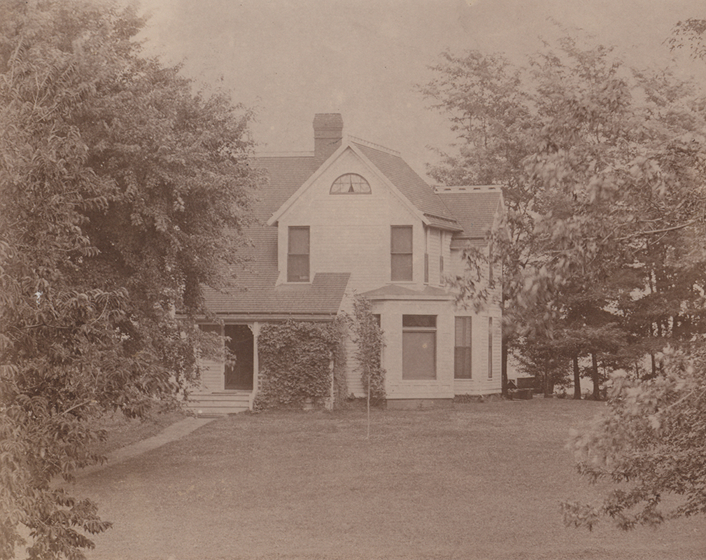 Photograph of Coburn House taken from the Knoll. It depicts a white two-story building with trees in the foreground. Ivy vines cover part of the front of the house.