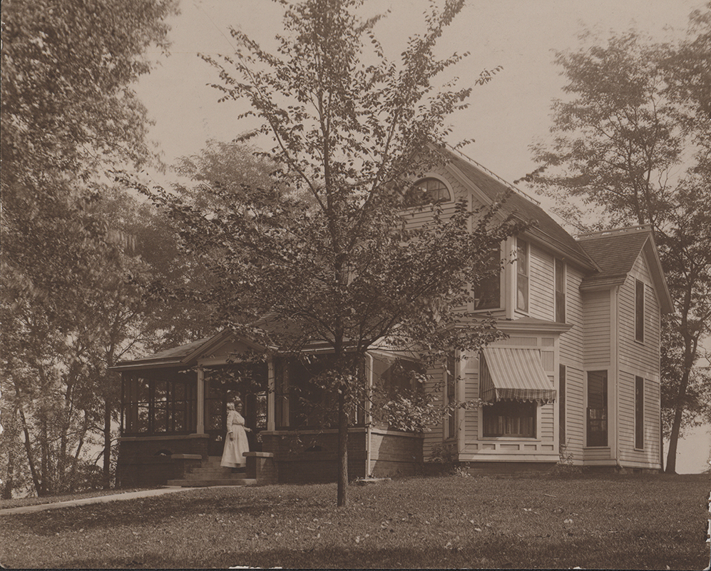 Photograph of Coburn House from the front. The front porch is screened in and a woman in a white dress stands on the front steps. The right front window has an exterior striped awning.