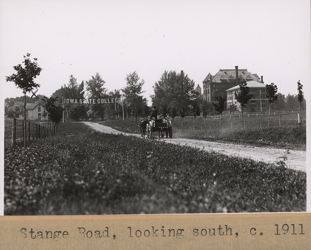 Photograph of Stange Road looking to the south. A man drives a horse-drawn cart towards the south. A sign reading Iowa State College crosses the road. Teamsters Cottage #2 is visible to the left. On the right, the campanile, Agricultural Hall, and the Engineer's Cottage are visible.