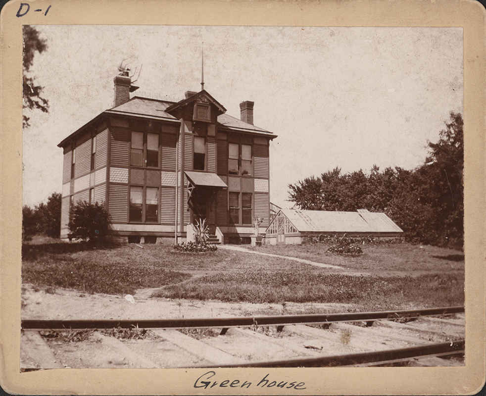Photograph of the first agricultural experiment station and its greenhouse. It is a two story building with an awning over the front stairs. A greenhouse sits in front of it and to the right. Train tracks are in the foreground.