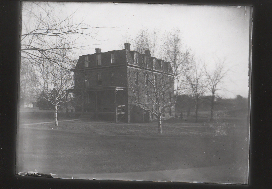 Photograph of the east boarding cottage, also known as Kirkwood Hall. The front and side of the building are shown. It is a brick building with a porch on the outside. Leafless trees surround the building.