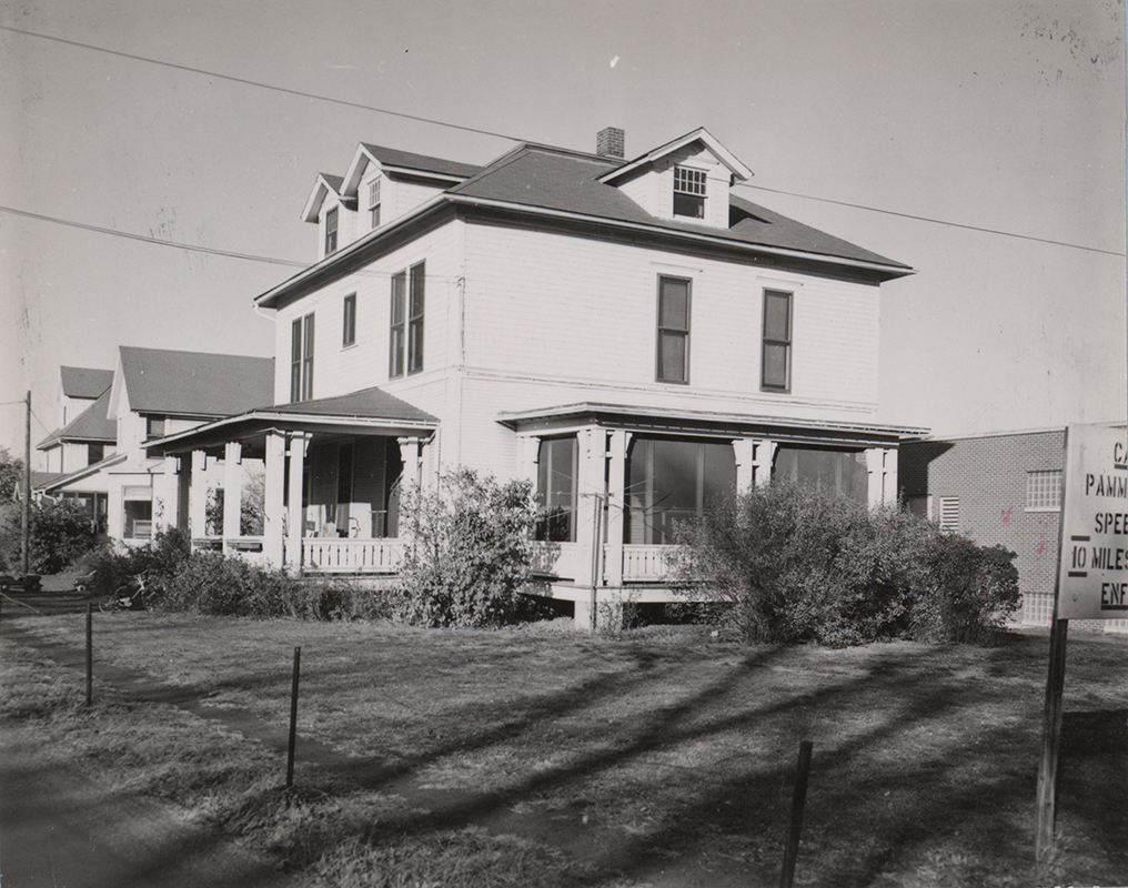 Photograph of Bevier House from the side. It has a screened in porch and a front porch. Two other houses are in the background. Part of a sign is visible in the foreground.