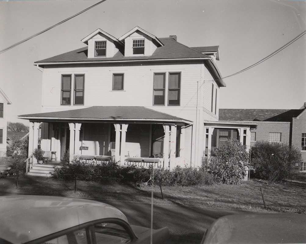 Photograph of Bevier House from the front. It is a white two-story house with a front porch. The top of two cars are in the foreground.