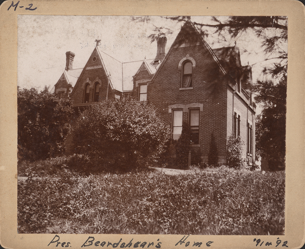 Photograph of The Gables, which was President Beardshear's house at the time. It later became the International House. The photograph shows a brick house with many visible gables. A large bush sits in front of the house.