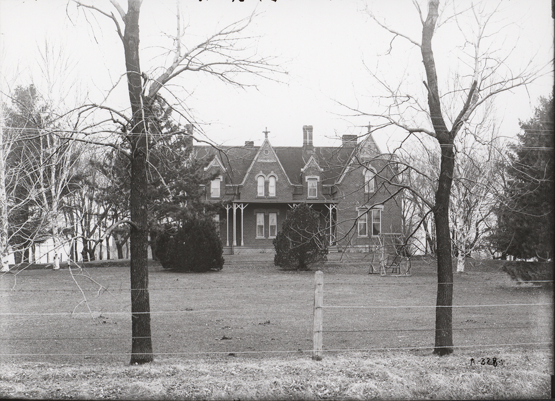 Photograph of the Gables in March, 1906. At the time it was the home of Professor Millikan Stalker. The photograph shows the front of the house with a porch. A seated swing is in the yard in front of the house. In the foreground are leafless trees and a barbed wire fence.
