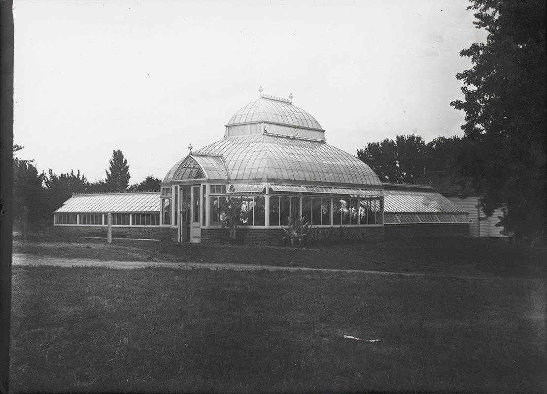 Photograph of a greenhouse on campus. It has a brick base and a barrel vaulted transept top. The caption reads, "Frame potting house where Horticulture Lab stood later from 1901-1978 on this spotl. Potting house moved to north of greenhouse."