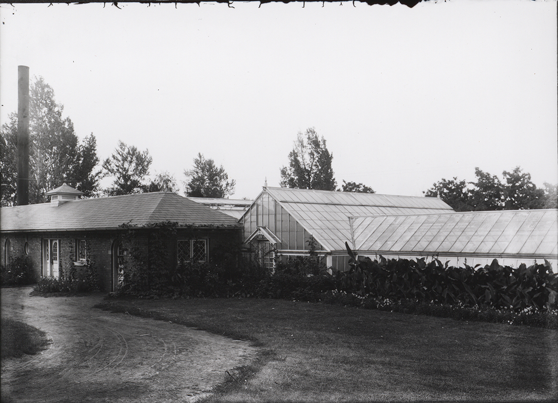 Photograph of a greenhouse on campus. It is long with an angled roof. A brick building sits in front of it. It served at different times as a potting and storage shed and the Ceramic Studio.