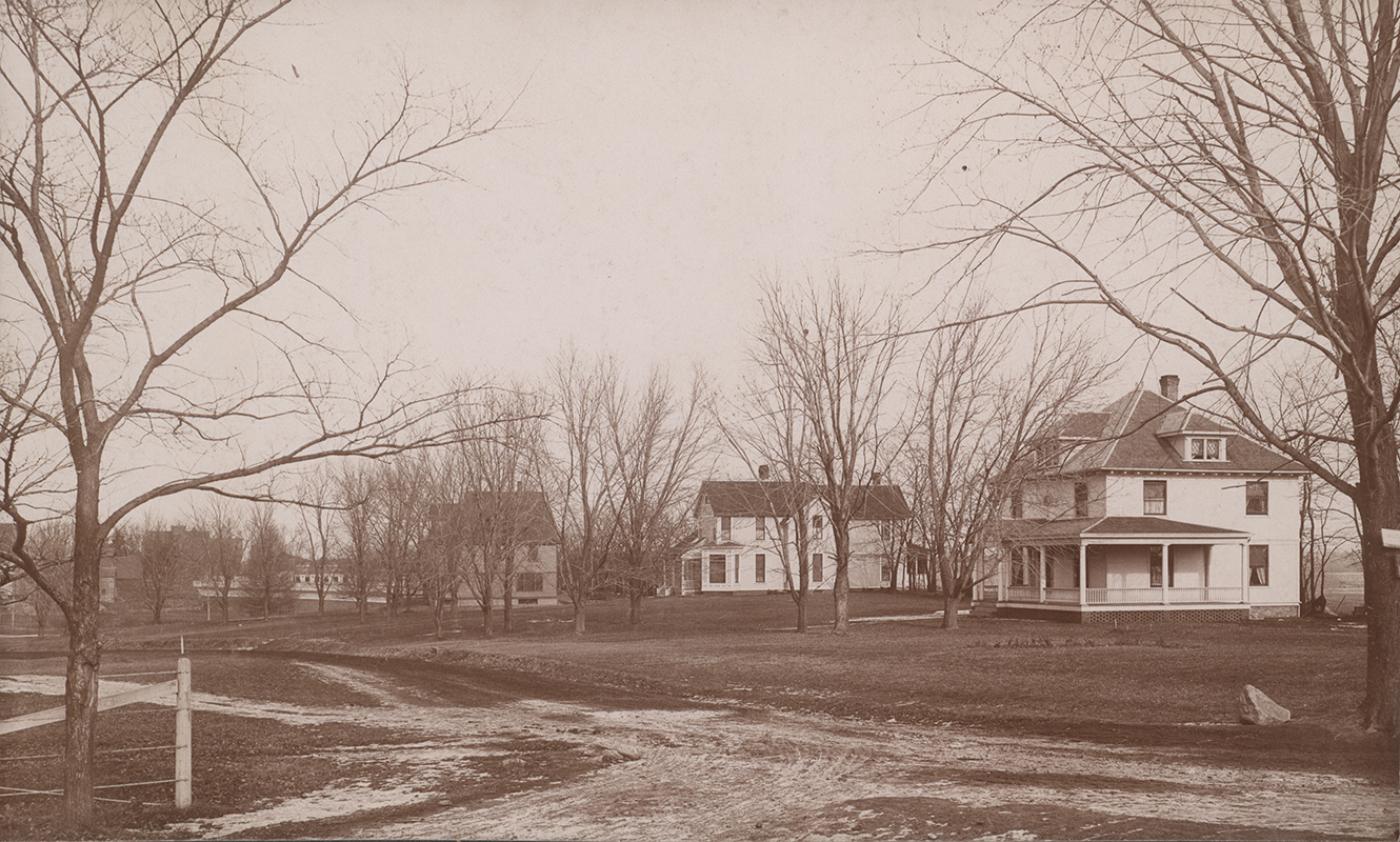Photograph of three houses on campus. From right to left: Kildee Cottage, Norton House, Coburn House. A dirt road is in the foreground and the houses are surrounded by leafless trees. The nearest two houses are white, and have front porches.