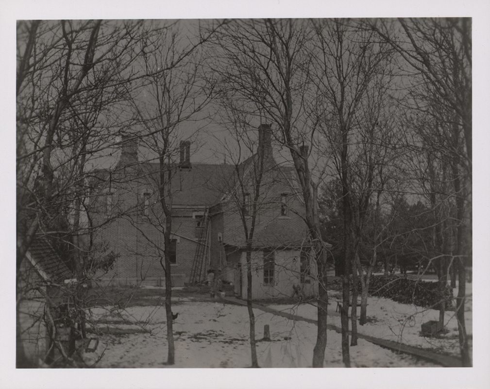 Photograph of the back of International House in winter. In the photograph, International House is in the snow, with leafless trees surrounding it. It is a brick building with an angled roof. Several windows are broken, and a ladder leans against the wall.